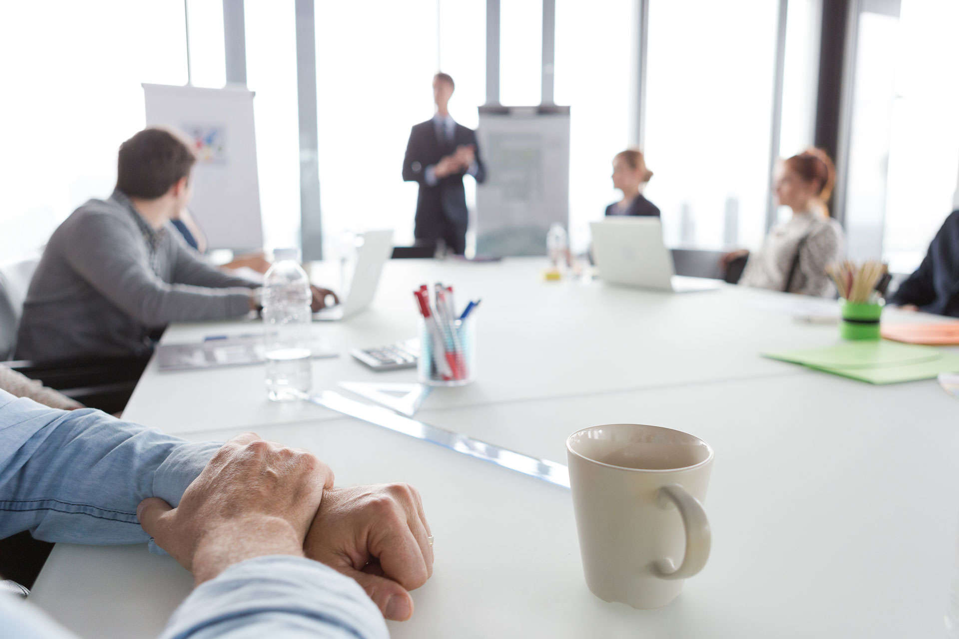 Multiple employees in professional attire sitting around a white conference table while watching a man give a presentation