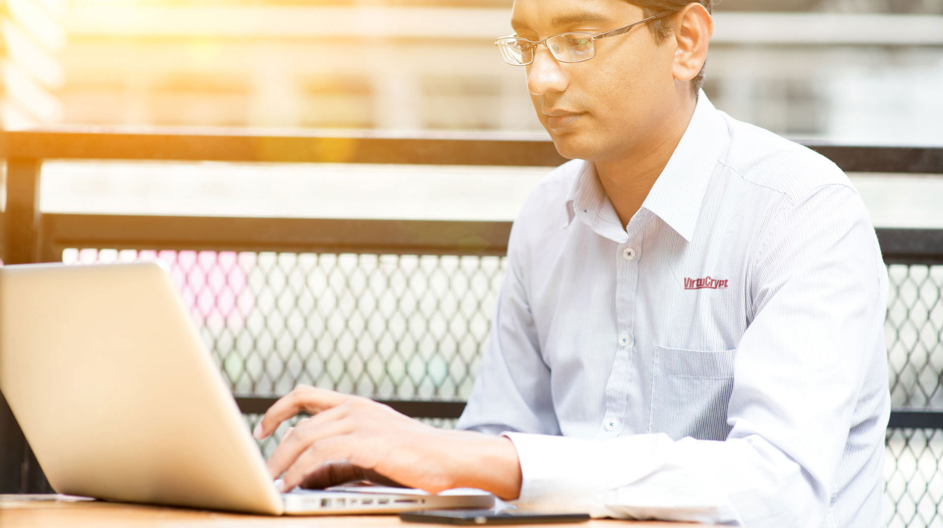 A man in a light-colored shirt in a sunny outdoor setting sitting down, facing a laptop, with his hand on the keyboard