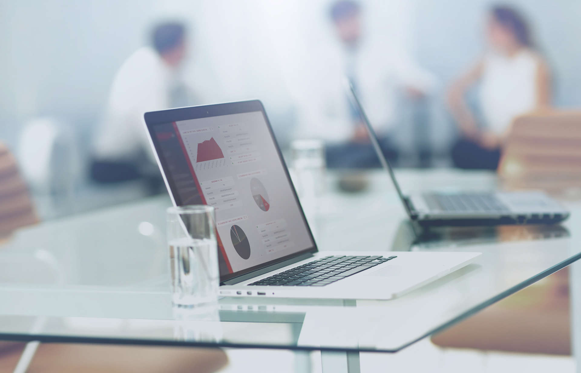 A Laptop featuring a VirtuCrypt Intelligence Portal GUI sits on a glass desk with three people sitting and talking in the background