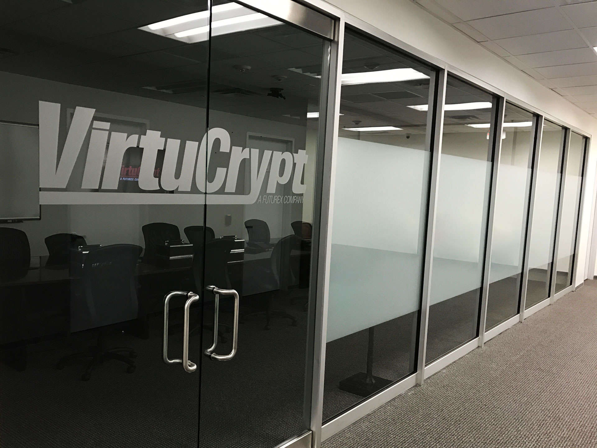 A wall of glass windows and a double glass door with a VirtuCrypt log leading to a conference room in a well-lit office