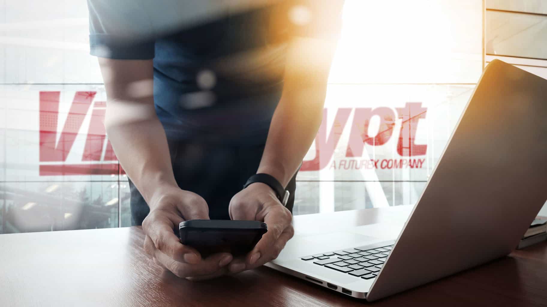 A man in a dark short-sleeved shirt leaning over a desk, holding a cell phone next to a laptop with sunlight coming in through a window in the background
